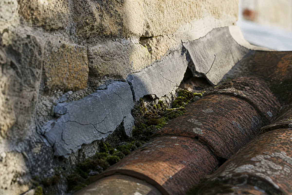Détail d’un solin ancien au mortier fissuré contre un mur en pierre, tuiles en terre cuite au premier plan